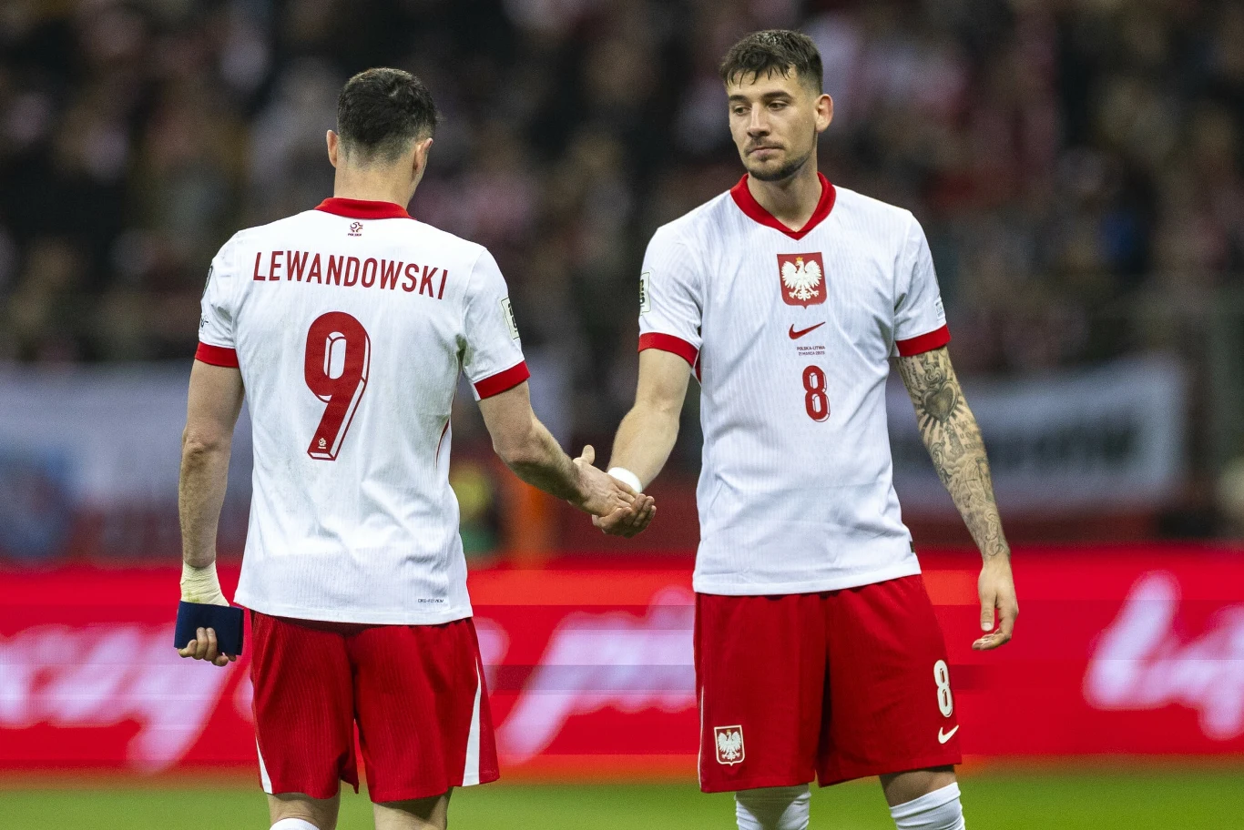 Robert Lewandowski i Jakub Moder Dwóch piłkarzy reprezentacji Polski w białych koszulkach z czerwonymi akcentami ściska sobie dłonie na stadionie, jeden z nich ma na plecach nazwisko Lewandowski i numer 9, drugi numer 8.