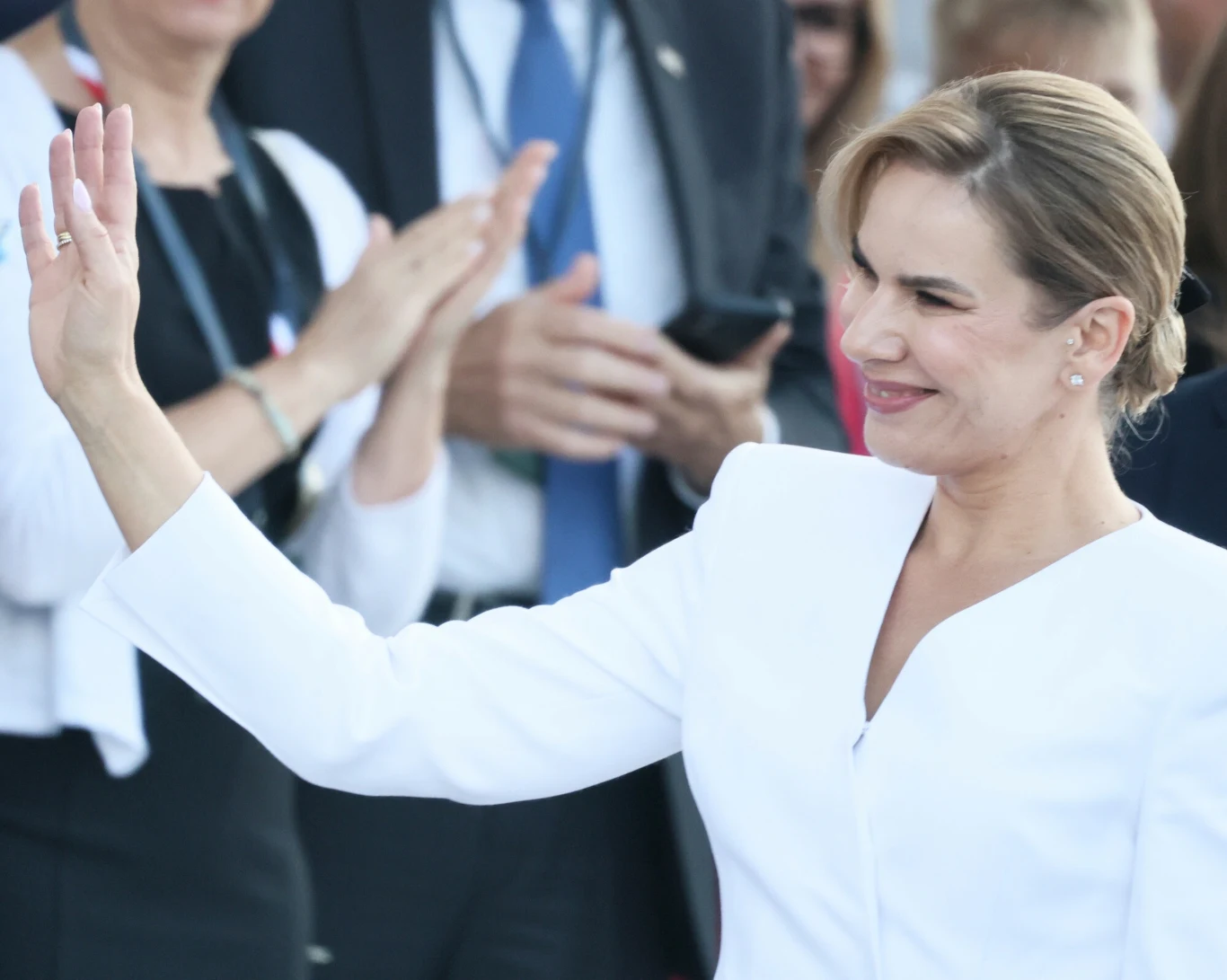 Uśmiechnięta kobieta w białym stroju unosi rękę w geście powitania lub pozdrowienia, w tle widoczne są inne osoby, niektóre z nich klaszczą.