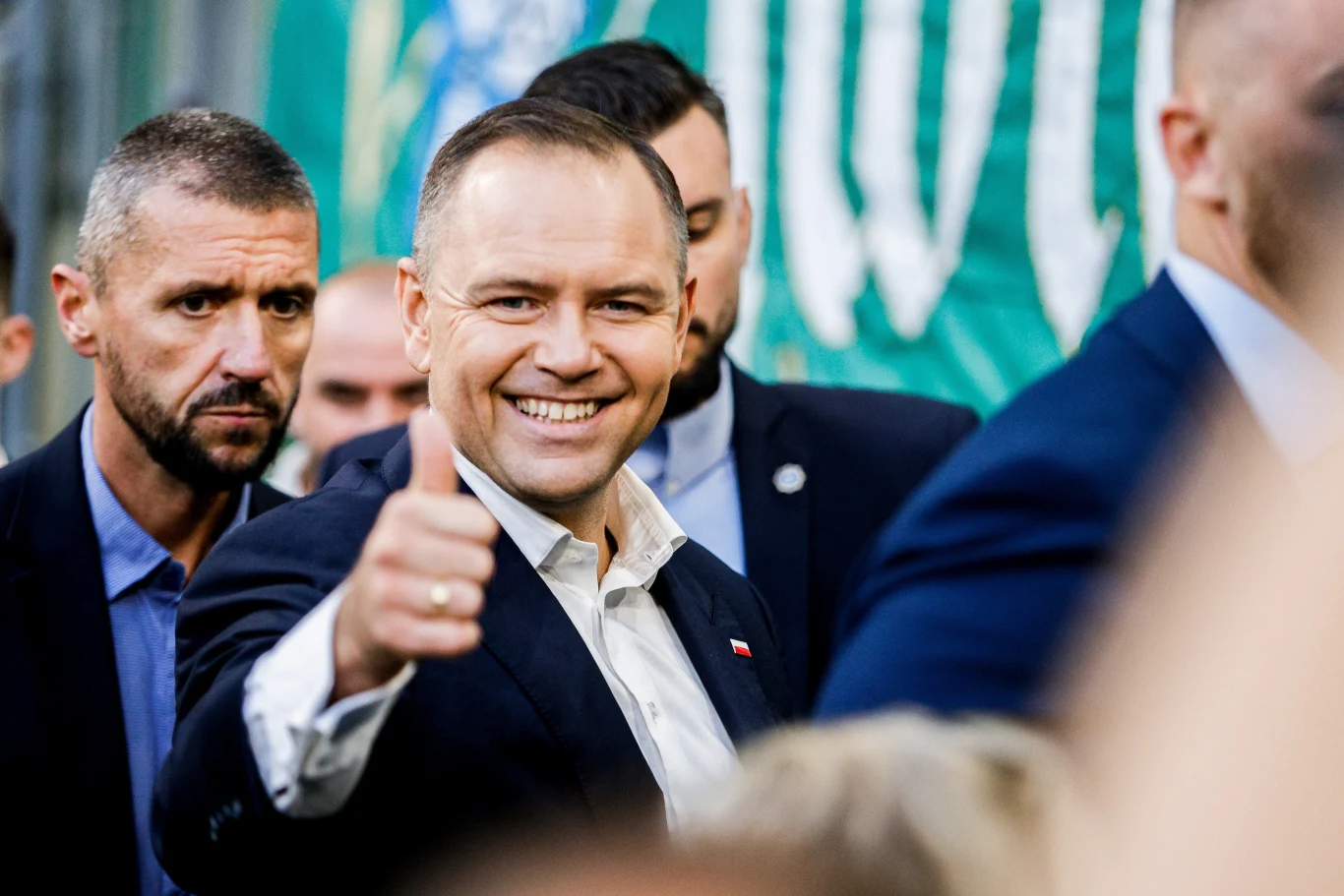 Mężczyzna w garniturze uśmiecha się i pokazuje kciuk uniesiony do góry, wokół niego stoją inni mężczyźni, a w tle widać zielony baner.