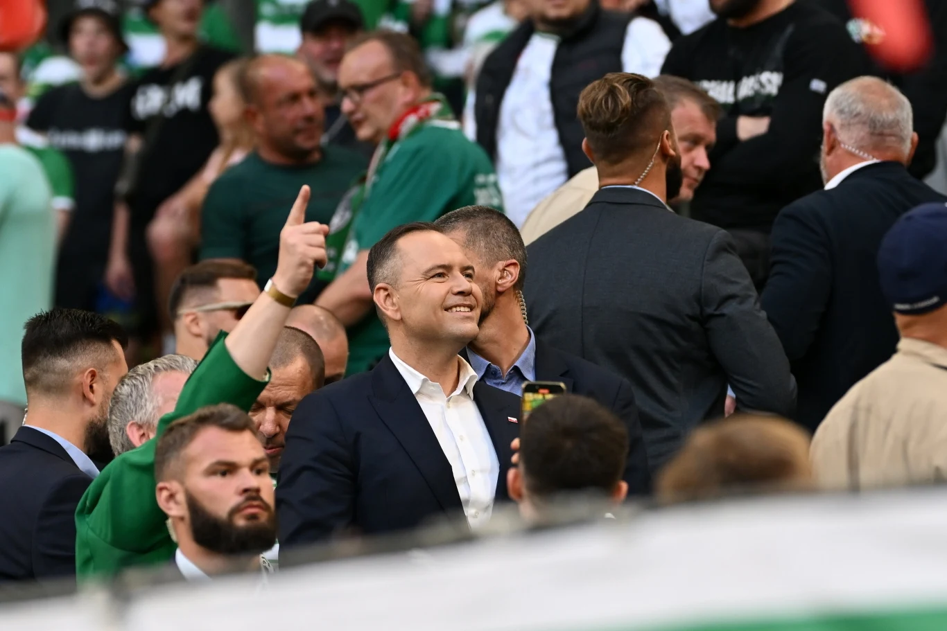 Karol Nawrocki na meczu Lechii Gdańsk Tłum ludzi na trybunach stadionu, w centrum kadru uśmiechnięty mężczyzna w garniturze, wokół niego inne osoby, w tle dominuje zieleń ubrań oraz otoczenia.