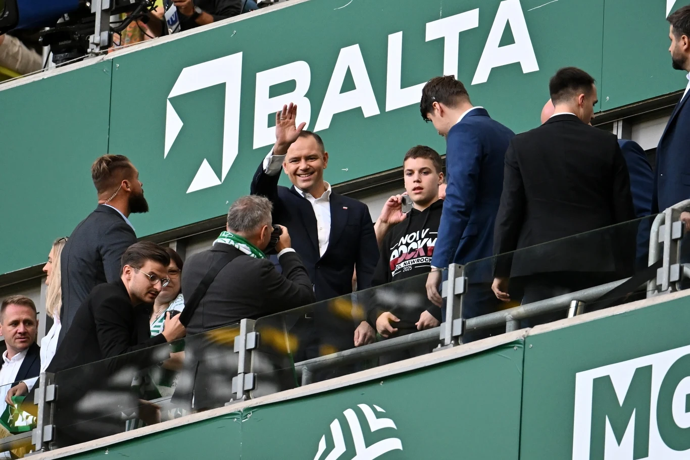 Grupa elegancko ubranych osób na trybunach stadionu, jedna osoba w środku kadru uśmiecha się i macha ręką, wokół niej stoją inni mężczyźni, a w tle widoczne logo firmy Balta.
