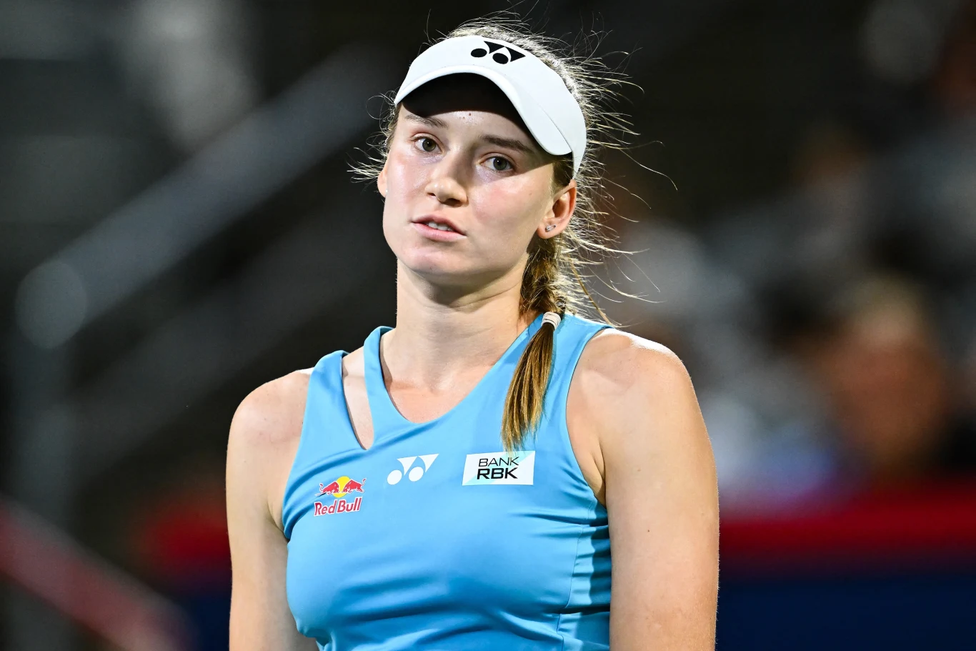 Jelena Rybakina Młoda kobieta w sportowym stroju tenisowym oraz białej czapce z daszkiem, na tle rozmytego stadionu, z neutralnym lub lekko zmęczonym wyrazem twarzy.