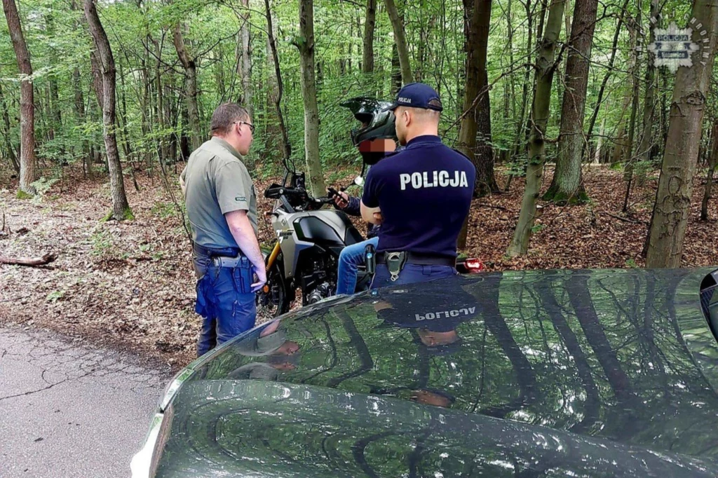 Stale rośnie w Polsce liczba quadów, motocykli oraz samochodów terenowych. Uszczelnienie przepisów jest niezbędne. Policjant oraz mężczyzna w stroju cywilnym rozmawiają z motocyklistą zaparkowanym przy lesie, na pierwszym planie fragment radiowozu.