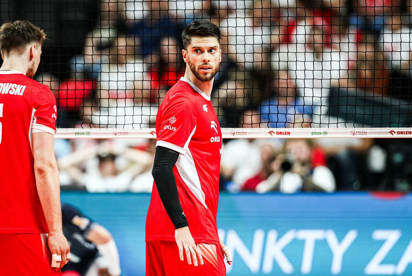 Aleksander Śliwka (w środku) w barwach reprezentacji Polski Siatkarz w czerwonym stroju z numerem na plecach stoi przy siatce podczas meczu, w tle widoczny tłum kibiców na trybunach.