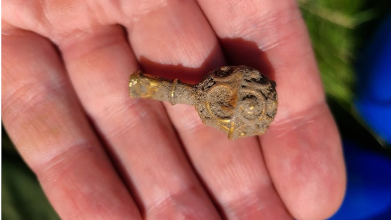 Dłoń trzyma niewielki, zardzewiały metalowy przedmiot o kulistym kształcie i zdobieniach, leżący na otwartej dłoni.