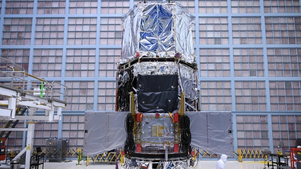 Duży satelita pokryty błyszczącą folią ochronną stoi we wnętrzu hali montażowej, wokół niego znajdują się różne elementy techniczne oraz osoba w kombinezonie ochronnym.