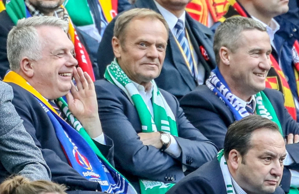 Donald Tusk na meczu Lechii Gdańsk w 2019 roku Mężczyźni w garniturach z szalikami klubowymi siedzą na trybunach stadionu podczas wydarzenia sportowego, uśmiechają się i żywo rozmawiają.