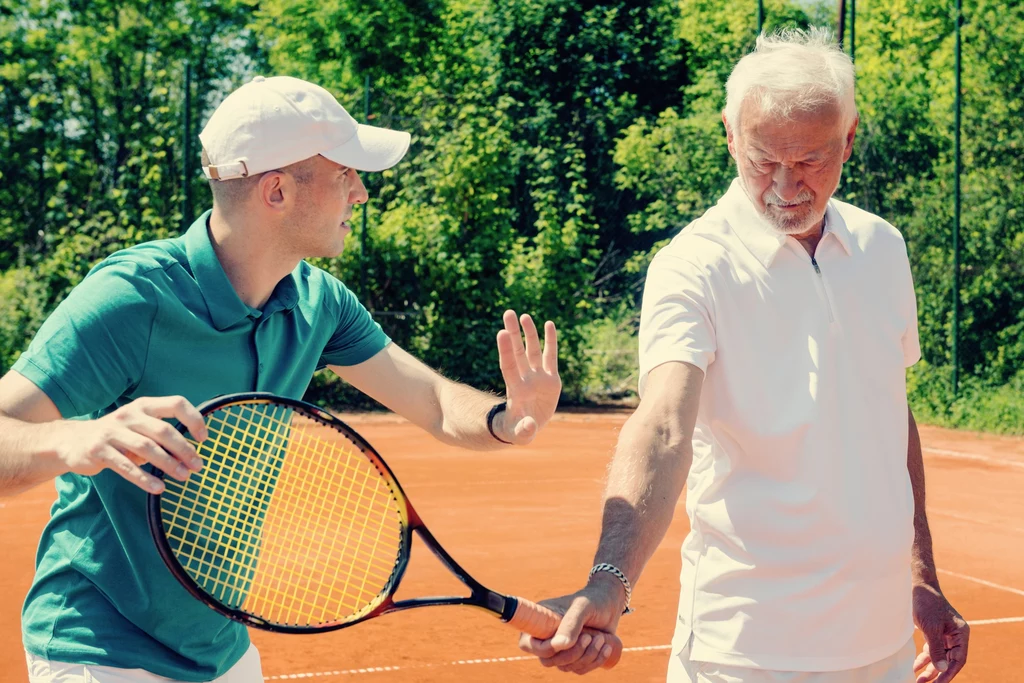 Młody mężczyzna uczy starszego mężczyznę trzymać rakietę tenisową na odkrytym korcie, otoczonym zielonymi drzewami; obaj ubrani w sportowe stroje.