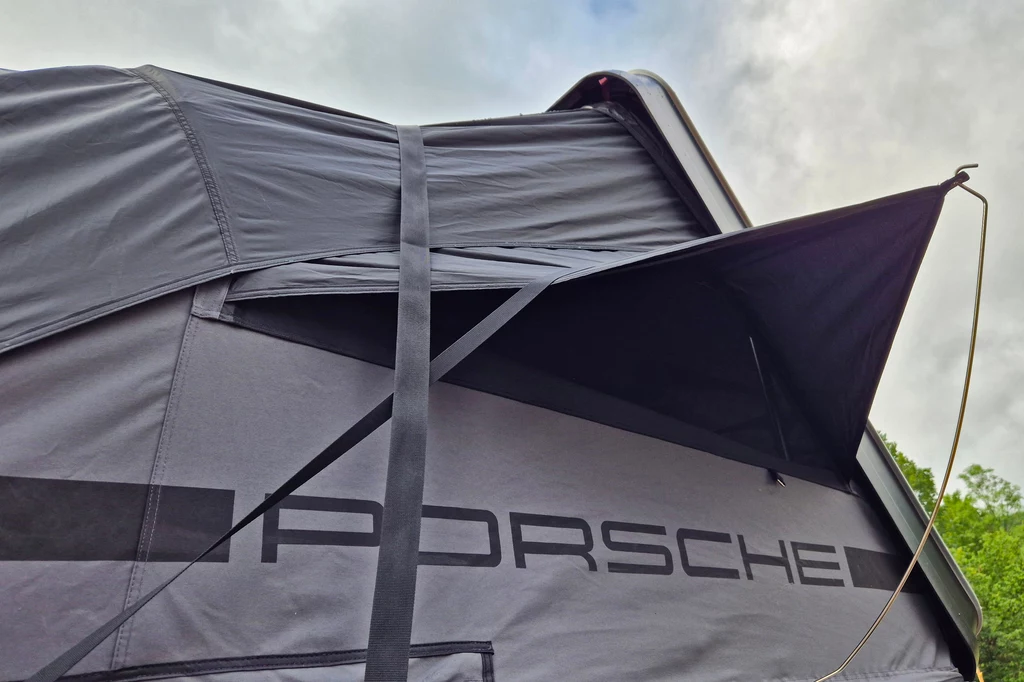 Czarny namiot dachowy z wyraźnym logo Porsche, rozłożonym na samochodzie, z widocznym fragmentem otwartego bocznego okna, na tle pochmurnego nieba.