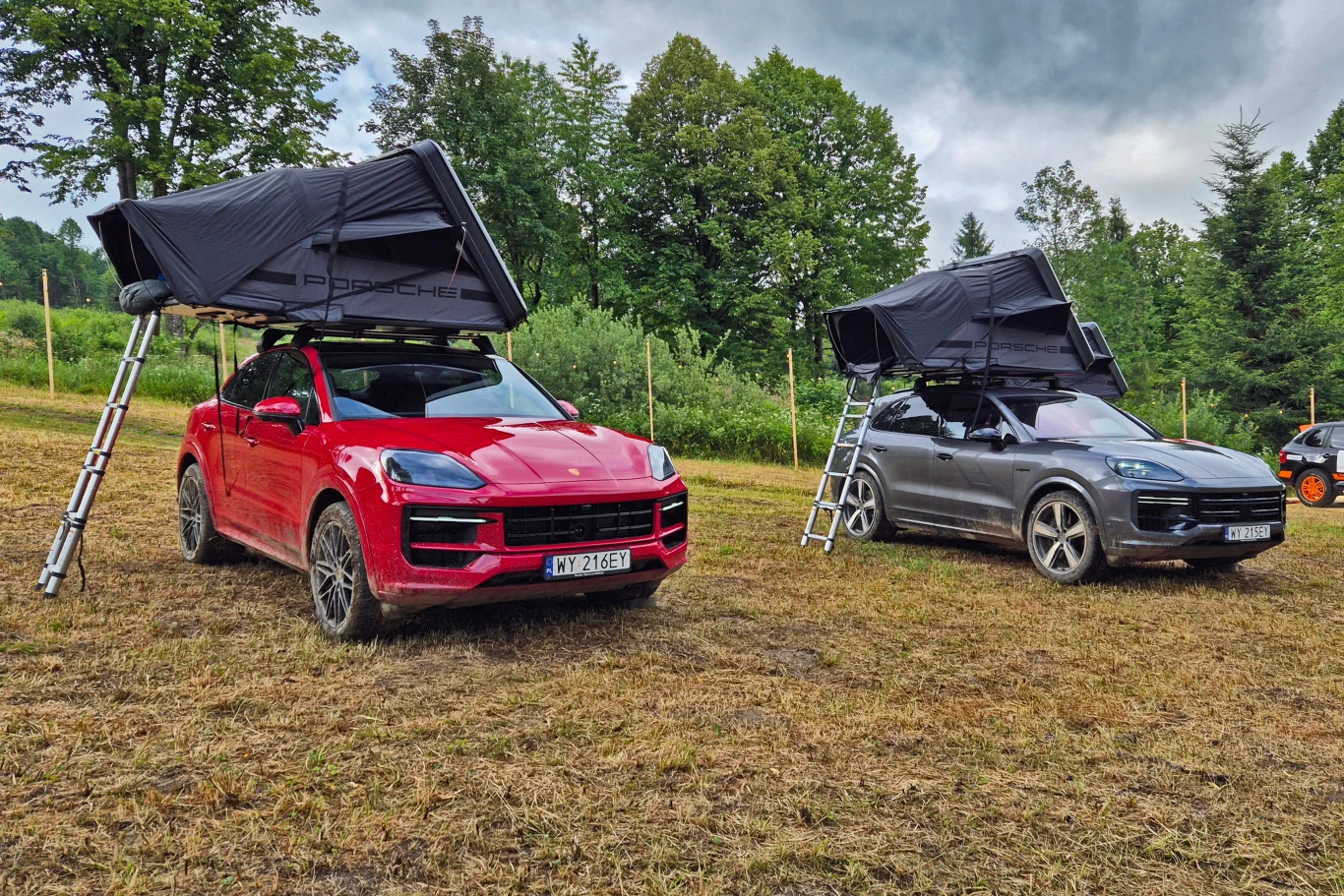 25 600 zł za namiot dachowy to niemało, ale najlepsze takie rozwiązania potrafią sporo kosztować i cena nie wynika tu z obecności napisu PORSCHE Dwa nowoczesne samochody terenowe z rozłożonymi namiotami dachowymi, ustawione na trawiastym terenie z otoczeniem gęstych zielonych drzew, niebo pochmurne, samochody stoją obok siebie, każdy z zamontowaną drabiną prowadzącą do namiotu.