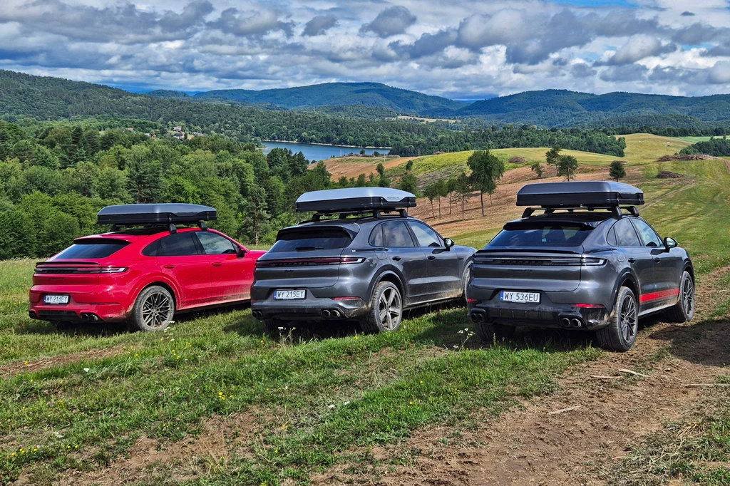 Trzy luksusowe SUV-y z boxami dachowymi zaparkowane na zielonym wzgórzu z widokiem na malownicze jezioro i pagórkowaty krajobraz w tle, otoczone letnią przyrodą i lasem.