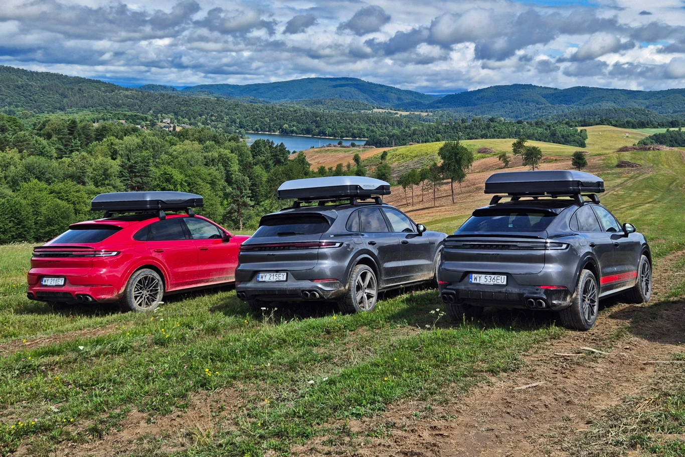 Góry, lasy, jeziora i wzgórza, które przemierzasz swoim SUV-em z namiotem dachowym - kusząca wizja, ale zwykle wymagająca odpowiedniego pozwolenia Trzy luksusowe SUV-y z boxami dachowymi zaparkowane na zielonym wzgórzu z widokiem na malownicze jezioro i pagórkowaty krajobraz w tle, otoczone letnią przyrodą i lasem.