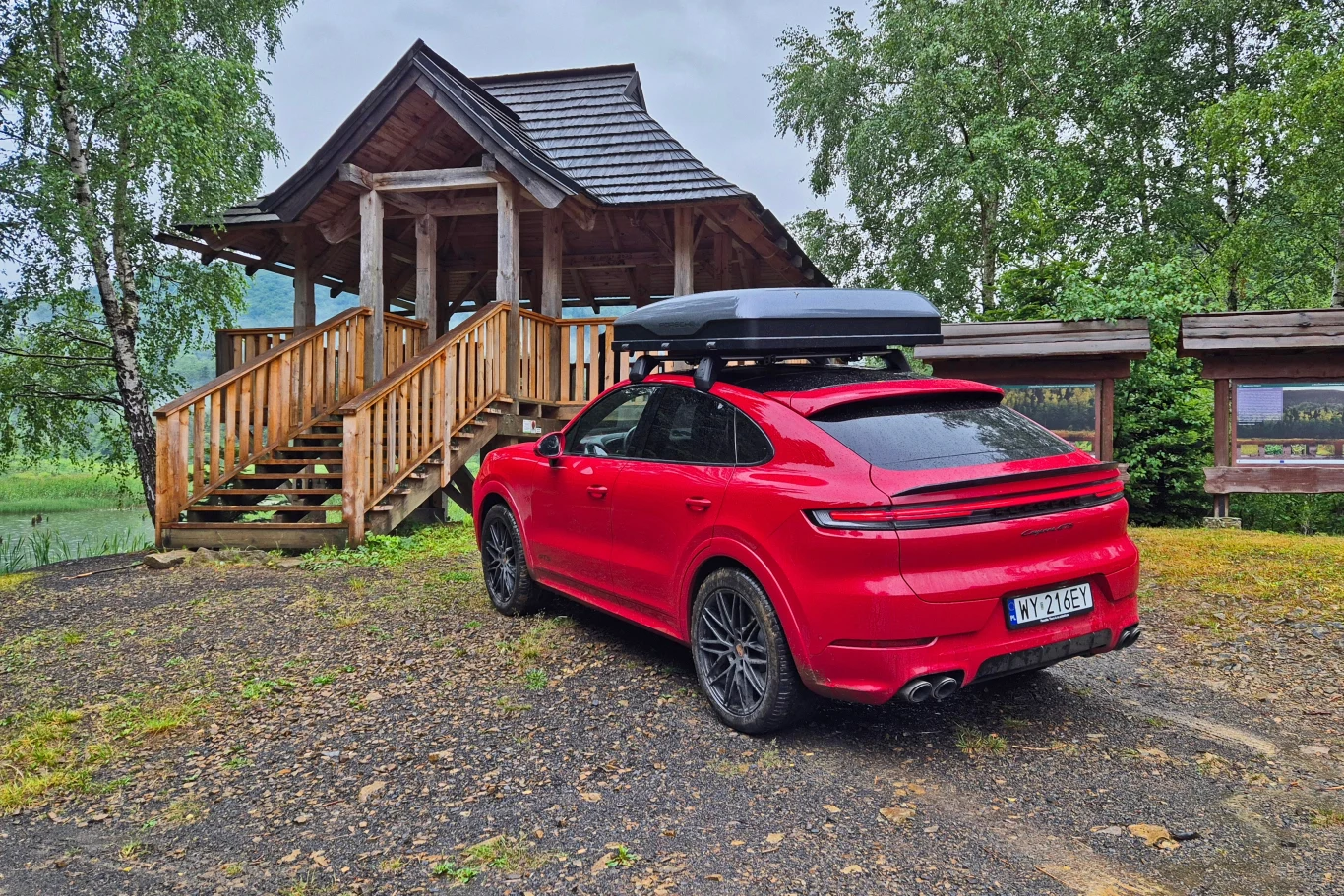 Porsche Cayenne z namiotem dachowym Czerwony samochód z boxem dachowym zaparkowany przy drewnianej altanie na tle zielonych drzew, podłoże pokryte żwirem oraz mokrą trawą.