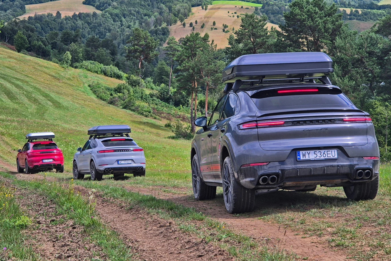 Porsche Cayenne z namiotem dachowym Trzy nowoczesne samochody terenowe z bagażnikami dachowymi jadą drogą polną przez zielone, pagórkowate tereny, otoczone lasem i wzgórzami.