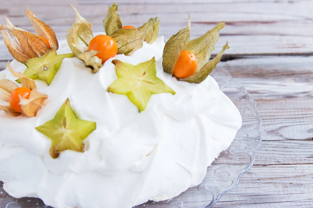 Przepisy z miechunką Beza z puszystym kremem, udekorowana plastrami karamboli oraz owocami physalis, ustawiona na przezroczystym talerzu na tle drewnianego stołu.