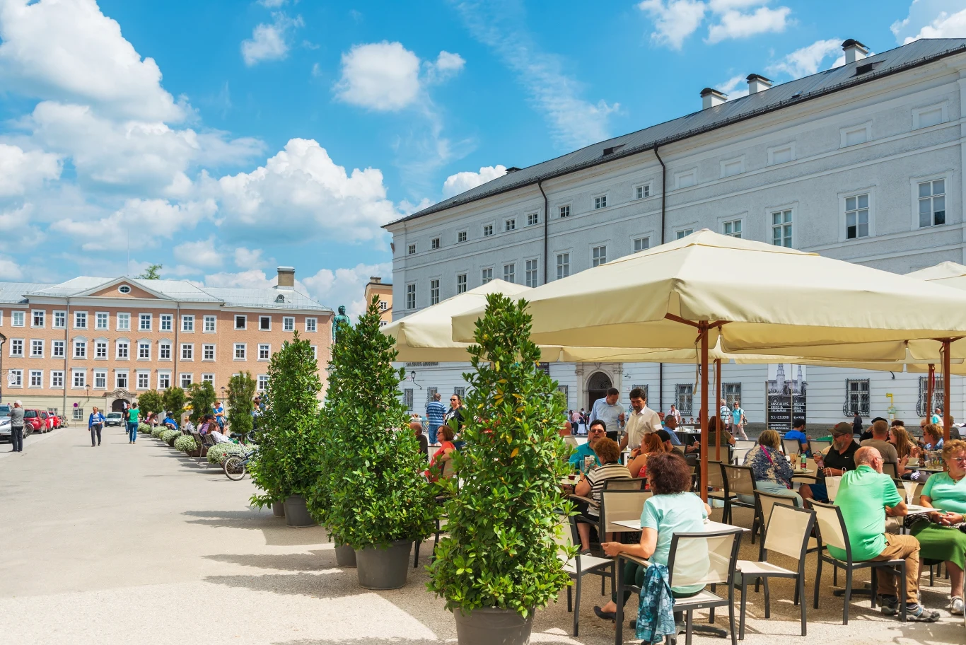 Restauracje w Austrii wprowadzają limity Ludzie siedzący przy stolikach pod parasolami w ogródku kawiarni na tle jasnych historycznych budynków, słoneczna pogoda, zieleń w dużych donicach oddzielająca strefę wypoczynku.