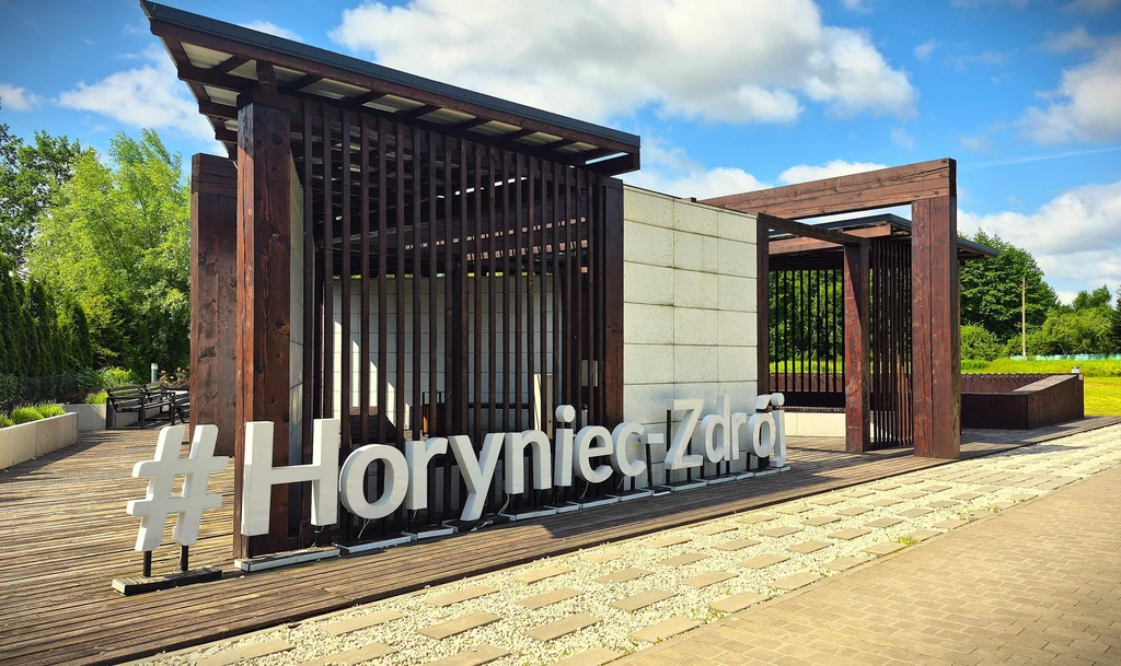 Nowoczesna konstrukcja architektoniczna z napisem #HoryniecZdrój wykonanym z dużych białych liter, otoczona zielenią i niebieskim niebem.