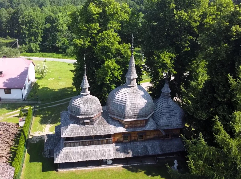 Drewniana cerkiew z trzema tradycyjnymi kopułami otoczona zielonymi drzewami, stojąca na zadbanym, zielonym trawniku obok białego budynku i ogrodzenia w wiejskim krajobrazie.