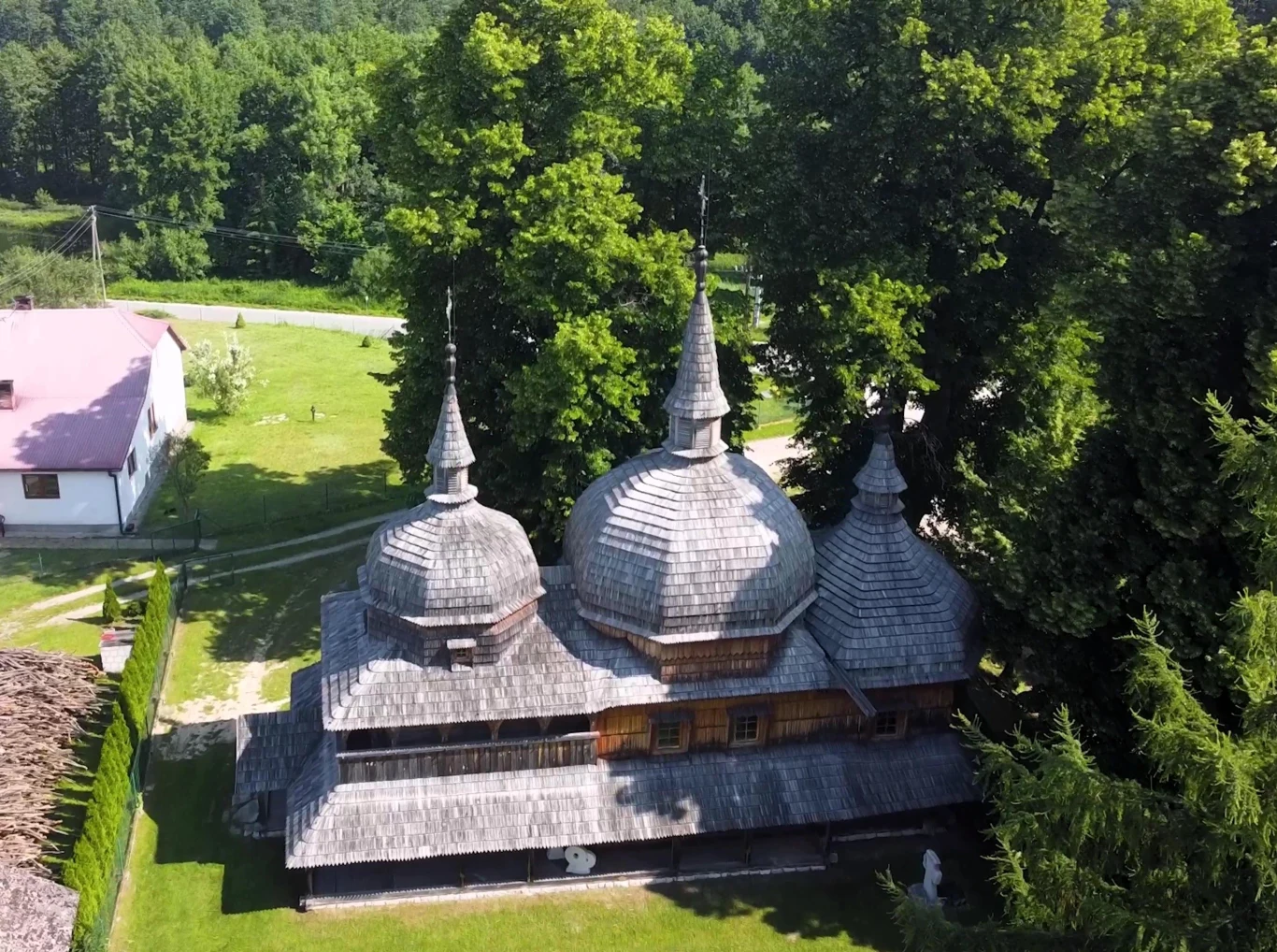 Drewniana cerkiew z trzema tradycyjnymi kopułami otoczona zielonymi drzewami, stojąca na zadbanym, zielonym trawniku obok białego budynku i ogrodzenia w wiejskim krajobrazie.