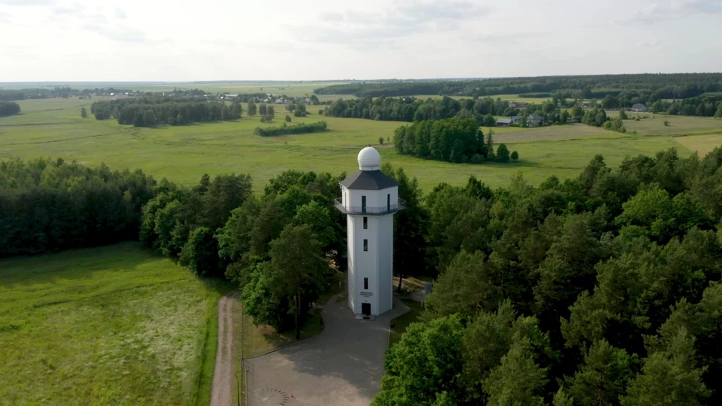Biała wieża obserwacyjna z kopułką na dachu otoczona gęstym lasem, w tle rozciągają się szerokie łąki i pola oraz pojedyncze zabudowania na horyzoncie.