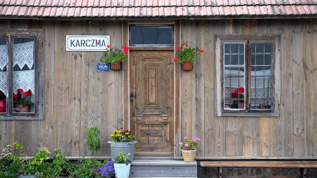 Drewniana fasada wiejskiej karczmy z centralnie umieszczonym wejściem, oknami po bokach ozdobionymi doniczkami z kwiatami oraz widocznym szyldem z napisem 'Karczma'.