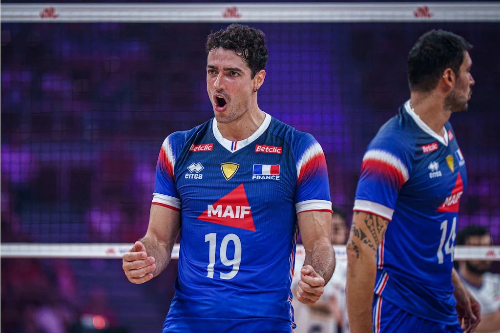 Yacine Louati (z numerem 19.) w barwach reprezentacji Francji Zawodnik reprezentacji Francji w siatkówce w niebieskim stroju sportowym okazuje emocje podczas meczu siatkarskiego, stojąc na boisku obok drugiego członka drużyny. Tło z niewyraźną publicznością podkreśla atmosferę sportowej rywalizacji.