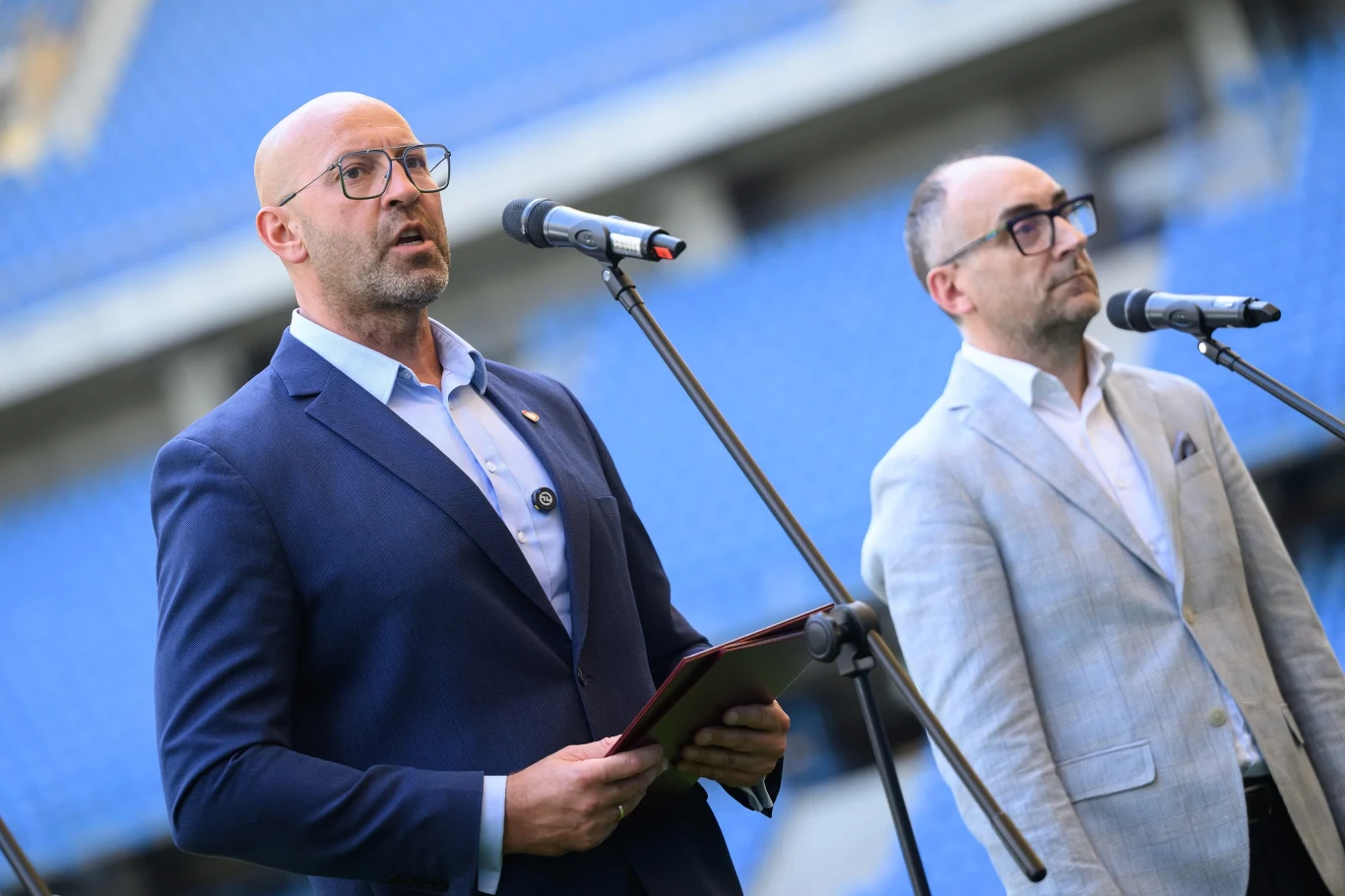 Na zdjęciu minister sportu i turystyki Jakub Rutnicki oraz prezes Lecha Poznań i członek zarządu PZPN Karol Klimczak podczas konferencji prasowej w Poznaniu Dwóch mężczyzn w garniturach przemawia do mikrofonów na tle niebieskich trybun stadionu, jeden z nich trzyma tablet.