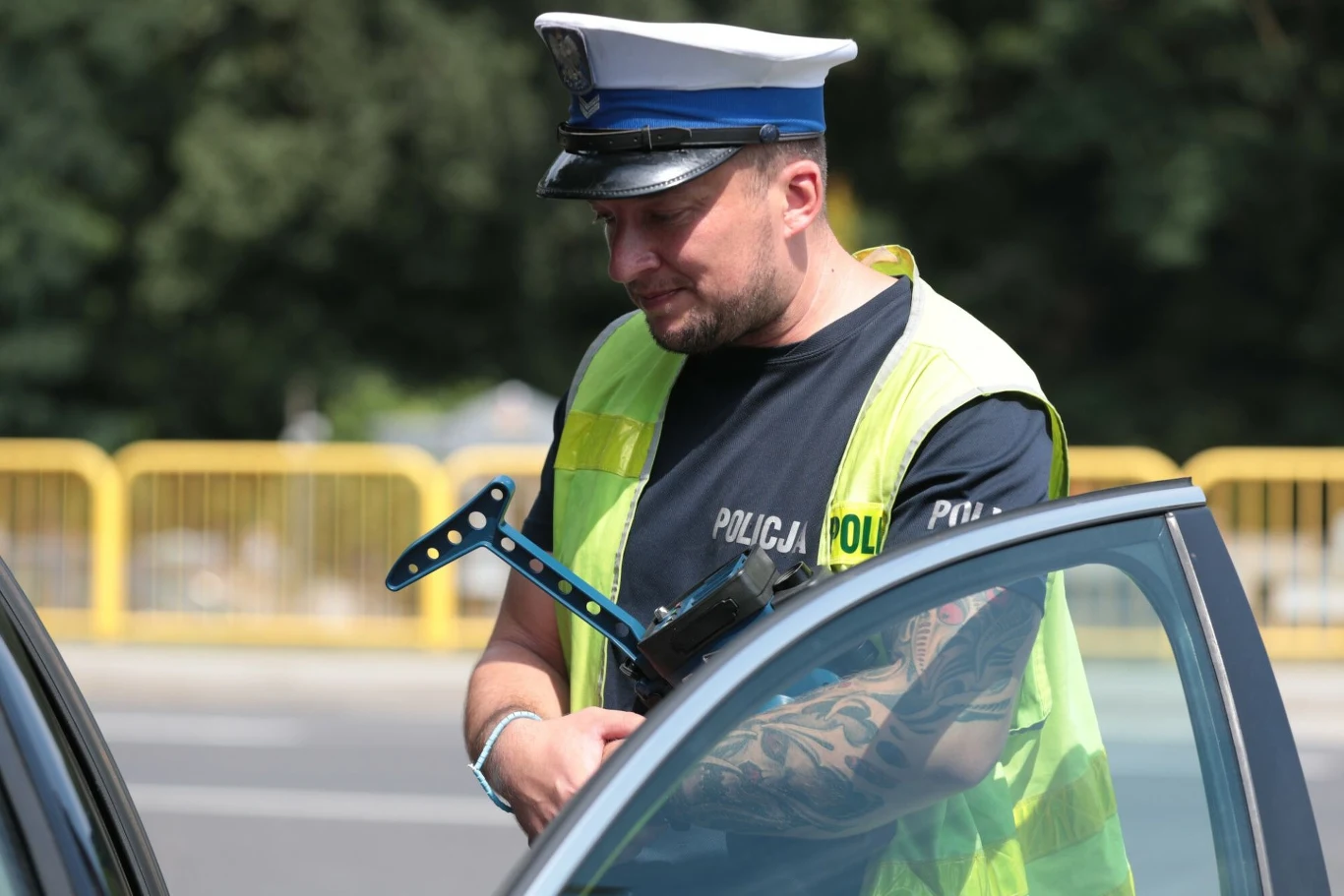 Rozpoczyna się akcja polskiej drogówki w ramach sojuszu Roadpol Policjant w czapce i kamizelce odblaskowej stoi przy otwartych drzwiach samochodu, w jednej ręce trzyma blokadę na koło auta, w tle widoczny żółty metalowy płot i rozmyte drzewa.
