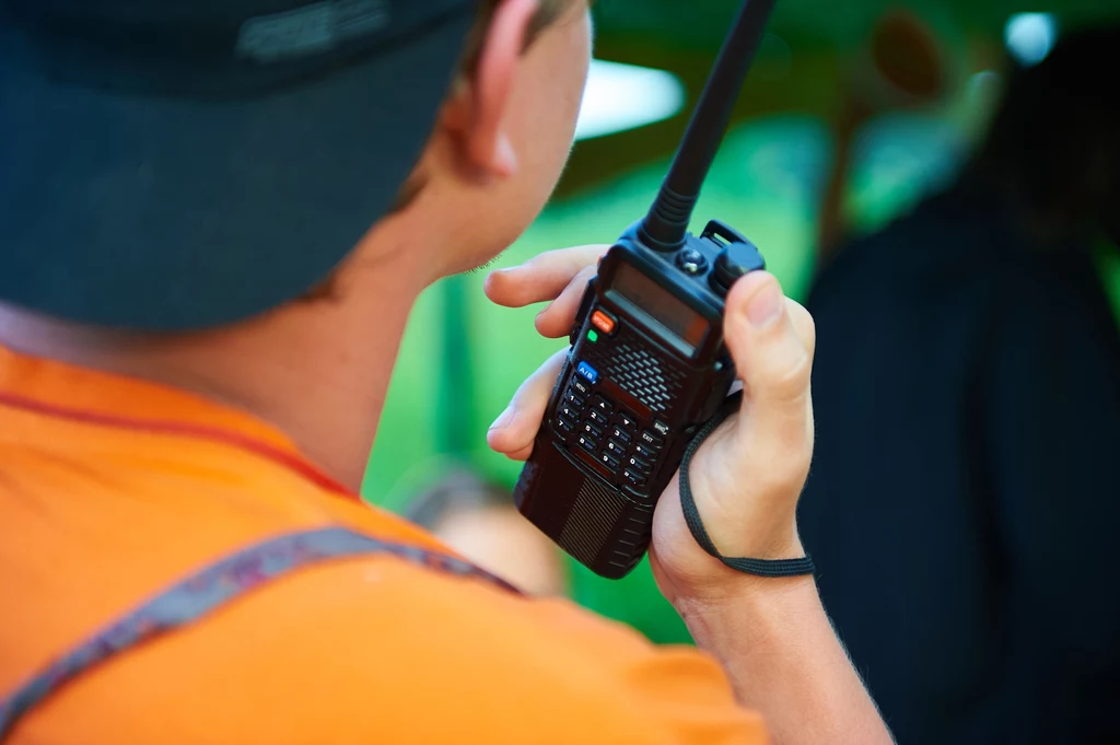 Osoba w pomarańczowej koszulce trzymająca radiotelefon, skupiona na rozmowie lub nasłuchiwaniu komunikatu, tło rozmyte w odcieniach zieleni.