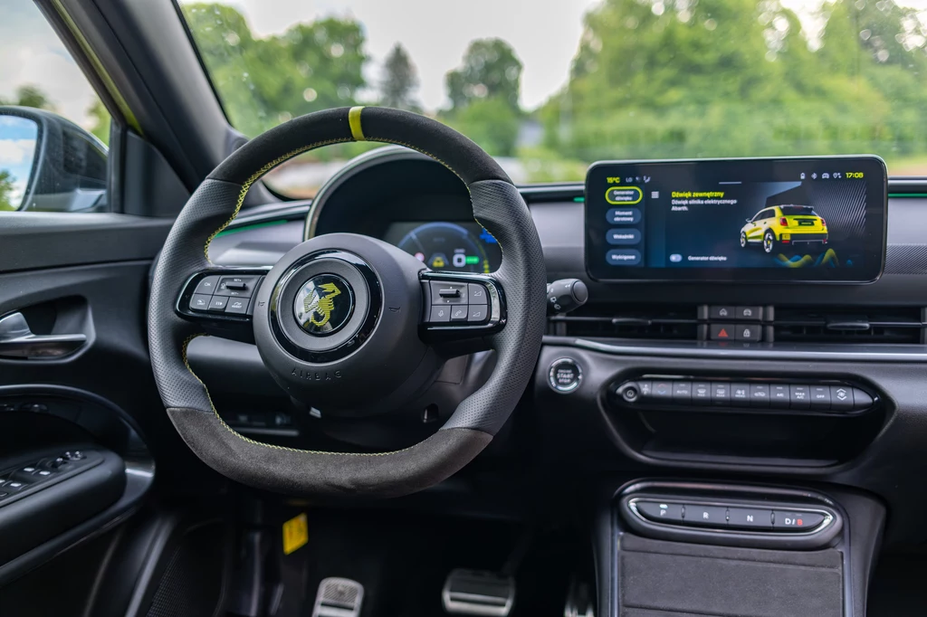 Jednym z najważniejszych wyróżników Abartha 600e, na tle bliźniaka z logo Fiata, jest sportowa kierownica pokryta skórą i alcantarą Kokpit nowoczesnego samochodu z kierownicą wykończoną skórą i żółtymi przeszyciami, dużym dotykowym ekranem wyświetlającym menu oraz cyfrowymi wskaźnikami, widoczna deska rozdzielcza i pedały, za oknem zieleń.