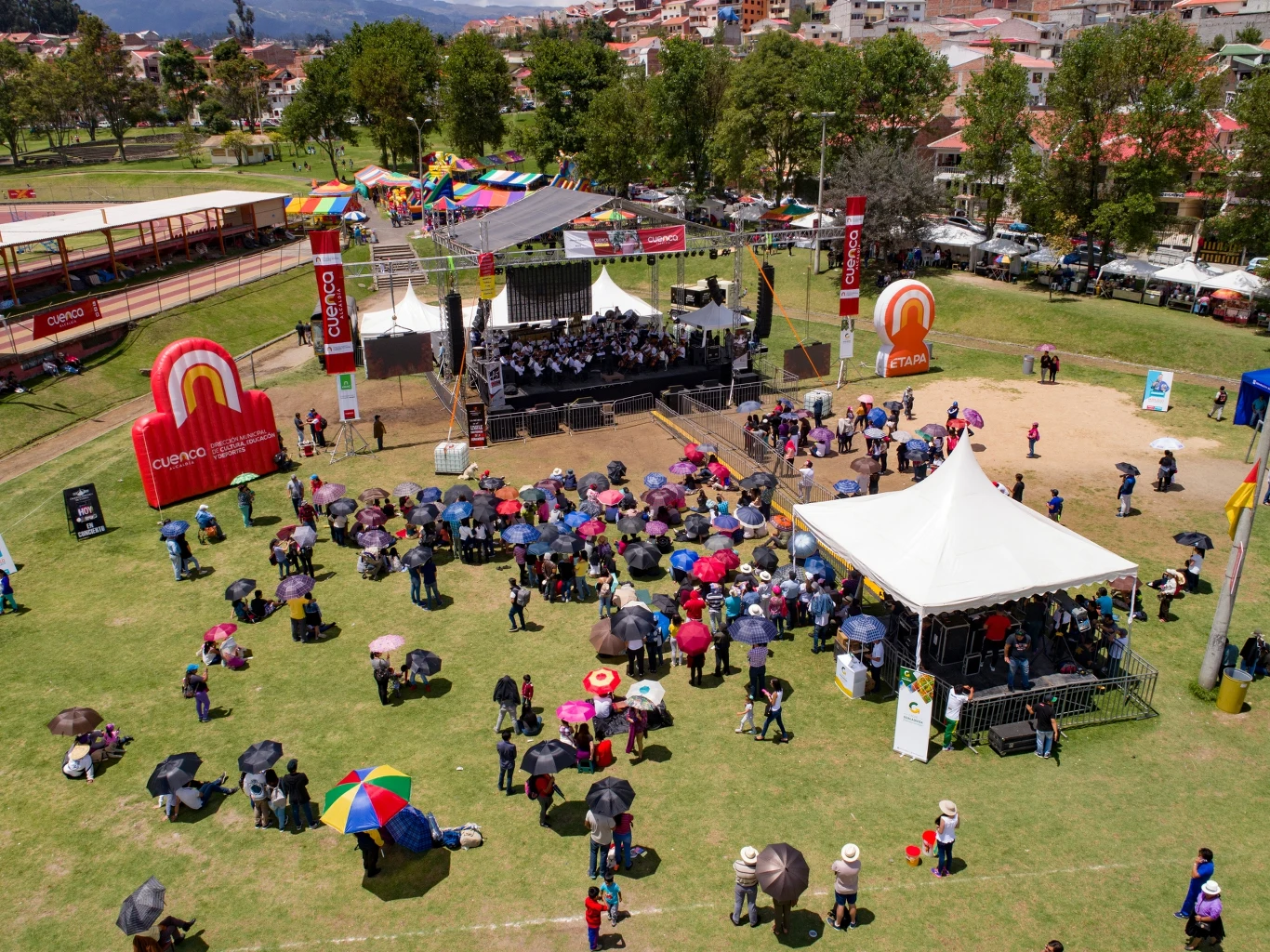 Grupa ludzi zgromadzona na trawiastym terenie parku uczestniczy w plenerowym wydarzeniu muzycznym, część osób trzyma parasole, na środku widoczna scena z zespołem muzycznym oraz duże namioty i kolorowe dekoracje.