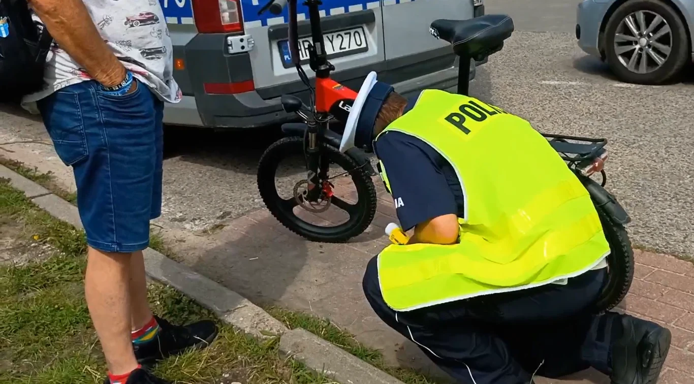Policja Mazowiecka skontrolowała rowery i hulajnogi Policjant w żółtej kamizelce odblaskowej kuca przy rowerze elektrycznym zaparkowanym koło policyjnego radiowozu, obok stoi mężczyzna w krótkich spodenkach i skarpetkach z kolorowymi paskami.
