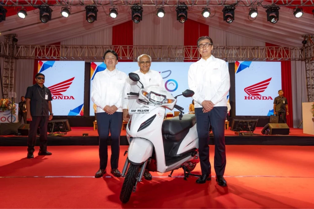 Trzech mężczyzn ubranych w białe koszule stoi na czerwonym dywanie przed białym skuterem Honda, w tle scena z dużym logo Honda oraz telebimami i jasnym oświetleniem.