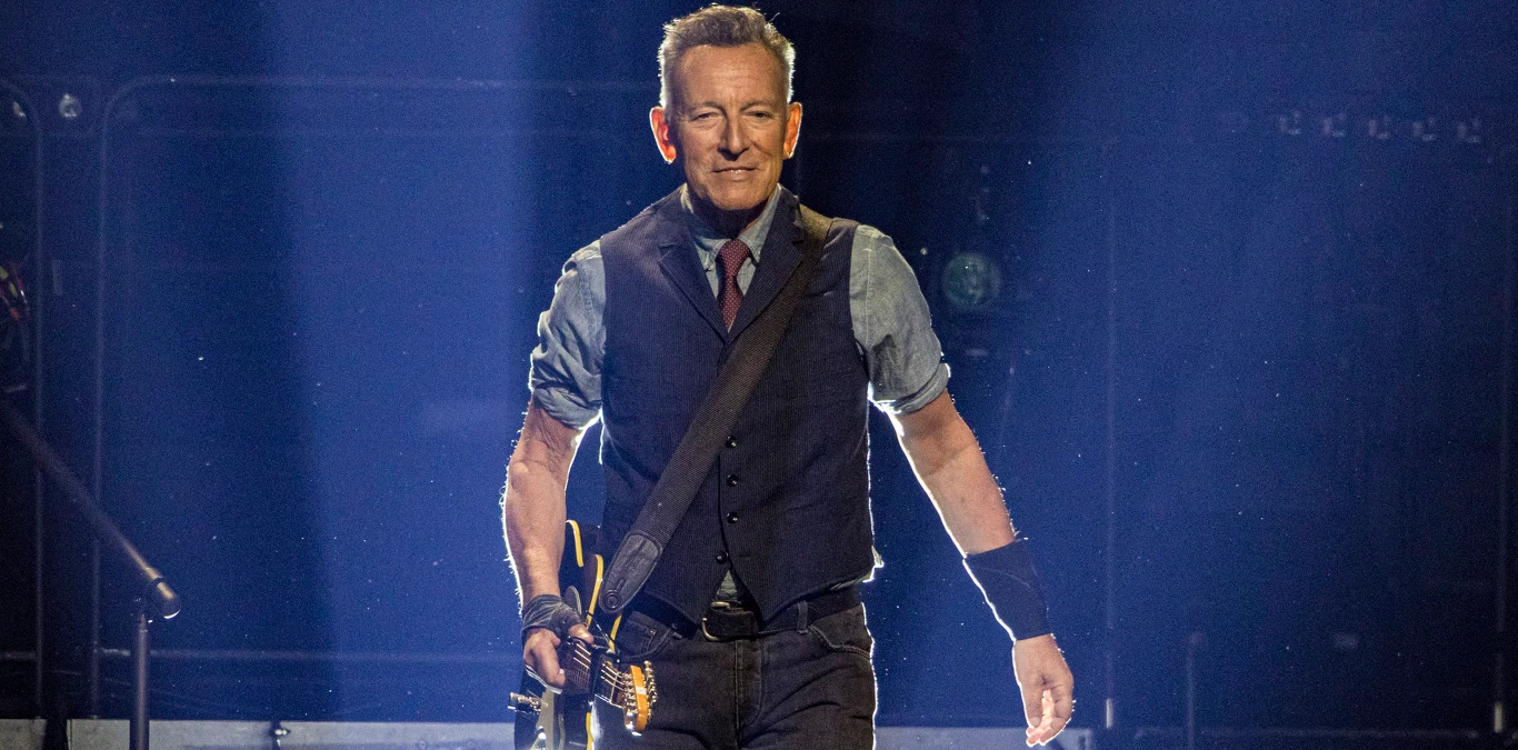 Bruce Springsteen na scenie w 2024 roku Mężczyzna w średnim wieku z gitarą przewieszoną przez ramię stoi na scenie w świetle reflektora, ubrany w koszulę, kamizelkę i krawat, z wyrazem skupienia na twarzy.