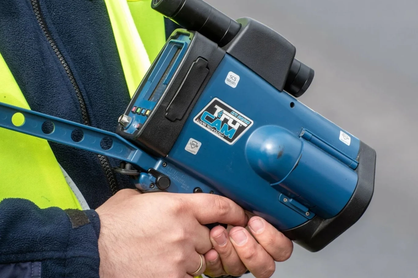 Nowe prawo wzbudza kontrowersje. Za złamanie grożą wysokie kary Osoba w kamizelce odblaskowej trzyma w dłoni niebieski radar do pomiaru prędkości, skierowany obiektywem na zewnątrz, na tle rozmytego, szarego tła.