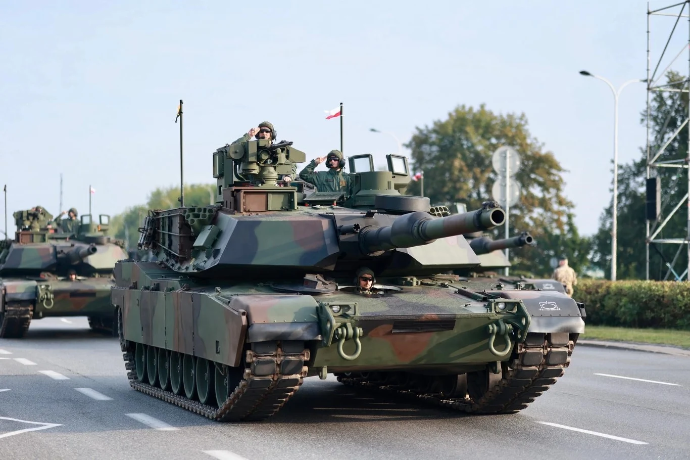 Czołgi palą dużo więcej od samochodów osobowych. Ale ile dokładnie? | Fot. Gov.pl Czołg wojskowy poruszający się po asfalcie, w tle drugi czołg, żołnierze znajdują się wewnątrz pojazdów, na czołgu widoczne są polskie flagi, otoczenie miejskie z drzewami i lampami ulicznymi.