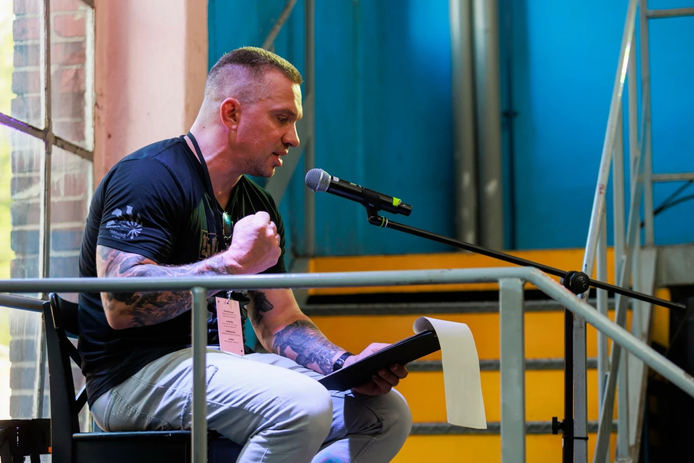 Jednym z gości cyklu Miejsca Literatury będzie Łukasz Orbitowski Mężczyzna z tatuażami na ramionach siedzi na krześle przy scenie i mówi do mikrofonu, trzymając zeszyt lub dokumenty oraz energicznie gestykulując pięścią, w tle metalowa konstrukcja i żółte schody.