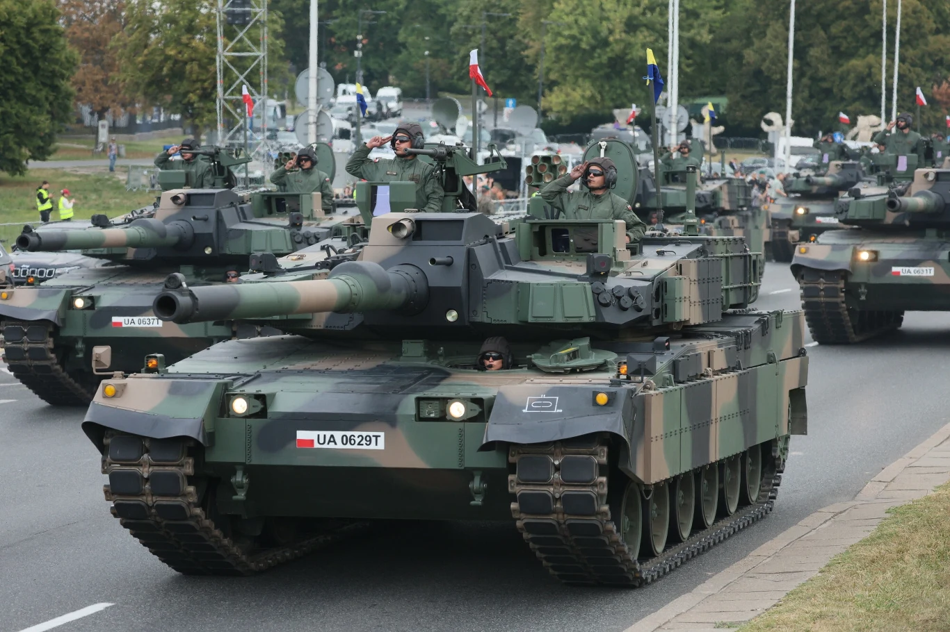 Koreański K2 Black Panther to jeden z najnowszych czołgów w polskiej armii Kolumna współczesnych czołgów wojskowych jadących po miejskiej ulicy, otoczonych przez żołnierzy w mundurach i pojazdy towarzyszące, widoczne flagi państwowe umieszczone na maszynach.