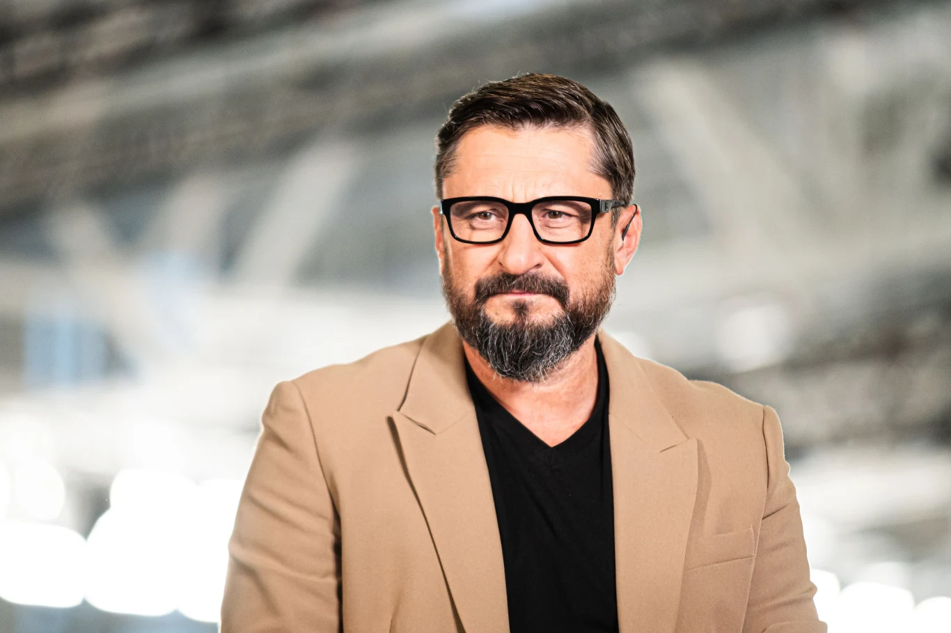 Mężczyzna w okularach i beżowej marynarce z brodą i krótkimi, ciemnymi włosami stoi na rozmytym tle przemysłowym lub biurowym. Wyraz twarzy poważny, a stylizacja elegancka, nowoczesna.