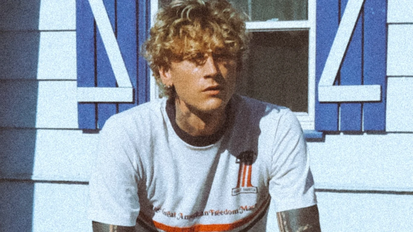Machine Gun Kelly Młody mężczyzna o kręconych blond włosach siedzący przed białym budynkiem z niebieskimi okiennicami, ubrany w białą koszulkę z nadrukiem, światło dzienne podkreśla rysy twarzy.