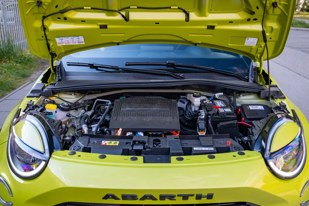 Abarth 600e dostępny jest w wersjach o mocy 240 KM oraz 280 KM Otwarta maska żółtego samochodu marki Abarth, wyeksponowany silnik oraz elementy komory silnikowej, widoczne reflektory i fragment ulicy w tle.