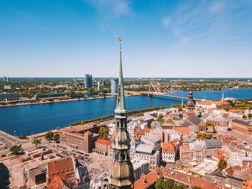Panorama miasta z wysoką wieżą kościelną na pierwszym planie, za którą rozciąga się widok na dachy historycznych budynków, rzekę z mostem i nowoczesną część miasta po drugiej stronie wody.