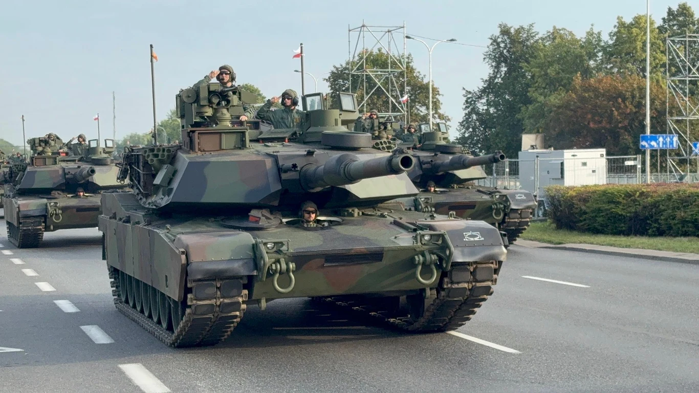 Czołgi wojskowe przemieszczają się kolumną po miejskiej drodze, żołnierze widoczni w wieżach pojazdów, wokół zabudowania miejskie i zieleń.