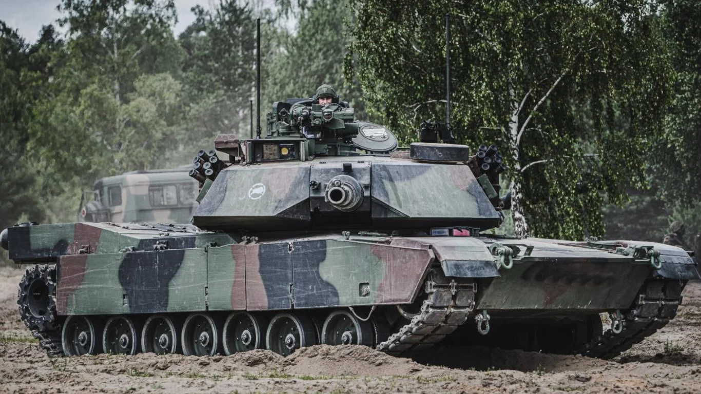 Czołg wojskowy w kamuflażu leśnym, stojący na ziemistym podłożu w otoczeniu drzew, z żołnierzem widocznym w wieżyczce oraz pojazdem wojskowym w tle.