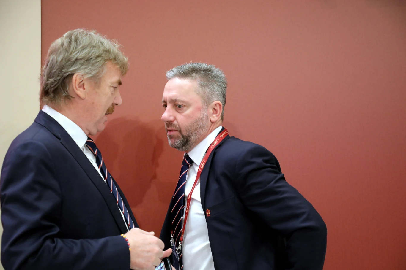 Dwóch mężczyzn w garniturach stojących blisko siebie, prowadzących poważną rozmowę przy czerwonej ścianie, obaj mają identyfikatory na szyjach.