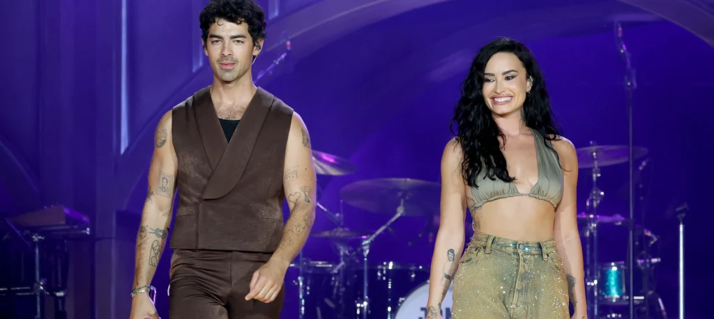 Dwie uśmiechnięte osoby, kobieta i mężczyzna, idą razem po scenie w jasnym oświetleniu, ubrani w modne stroje, w tle widoczne są elementy sceniczne i perkusja.