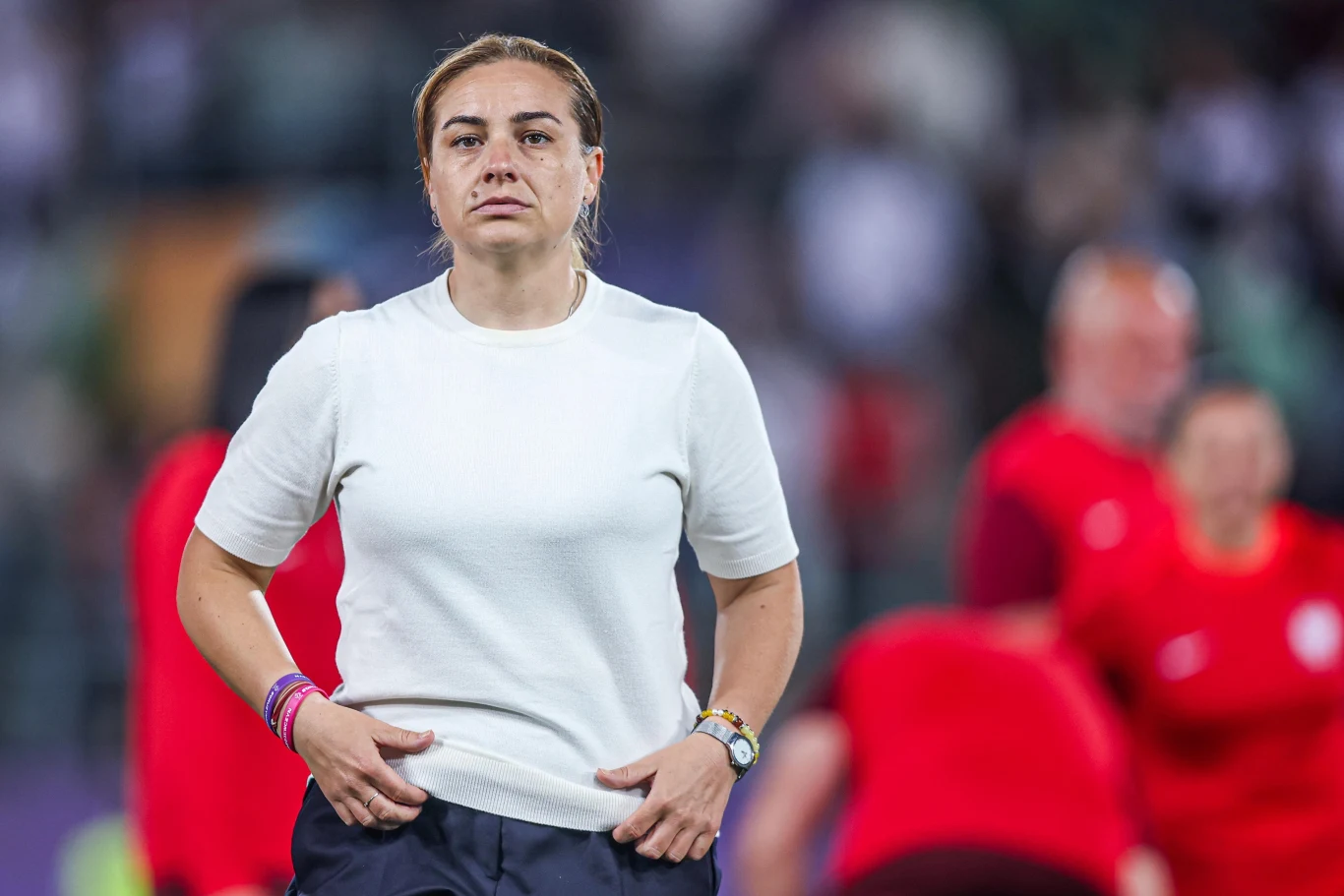 Nina Patalon Kobieta w białej koszulce i granatowych spodniach stoi w sportowym otoczeniu, z poważnym wyrazem twarzy. W tle widoczne osoby ubrane w czerwone stroje, rozmazane przez płytką głębię ostrości.