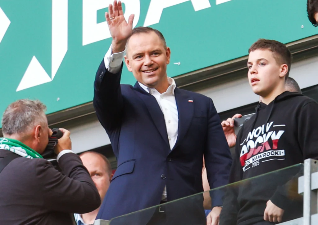 Uśmiechnięty mężczyzna w garniturze macha dłonią do tłumu, obok stoi nastolatek w czarnej bluzie, a po lewej mężczyzna z aparatem robi zdjęcie, w tle widoczny jest fragment zielonego baneru.