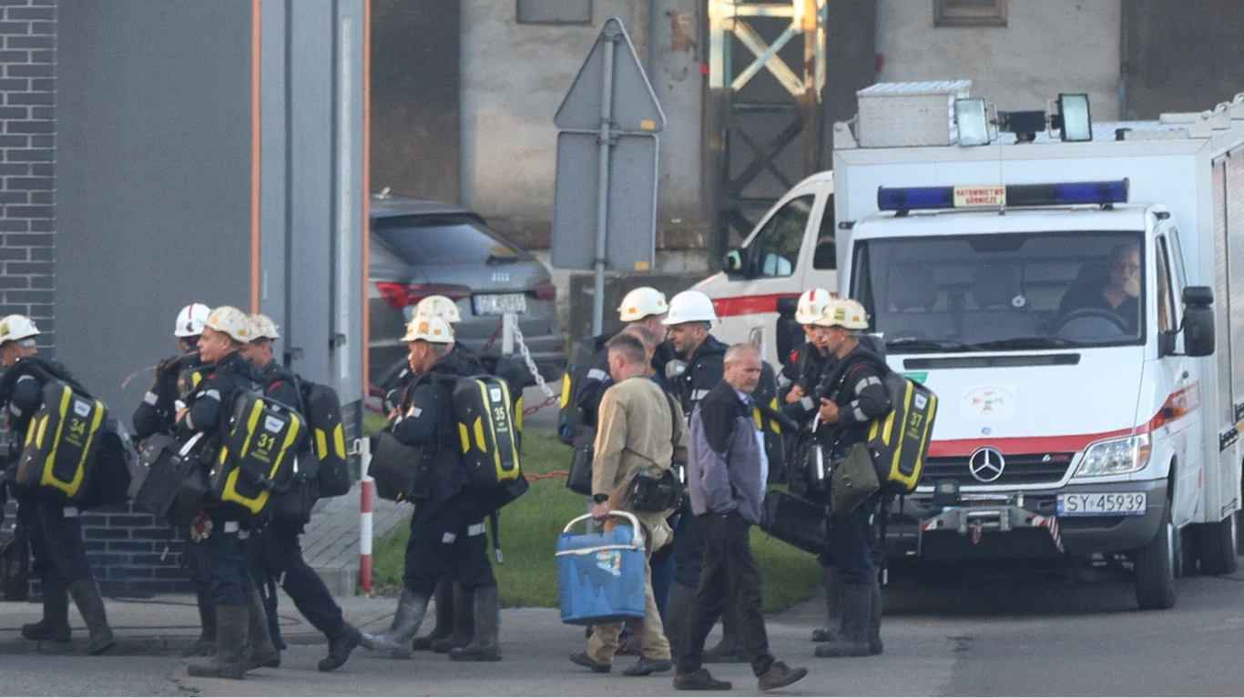 Poszukiwania górnika trwały od poniedziałku Grupa ratowników wyposażonych w aparaty tlenowe i kaski przemierza teren przemysłowy obok pojazdu ratowniczego, na pierwszym planie dwóch mężczyzn trzymających sprzęt ratowniczy.