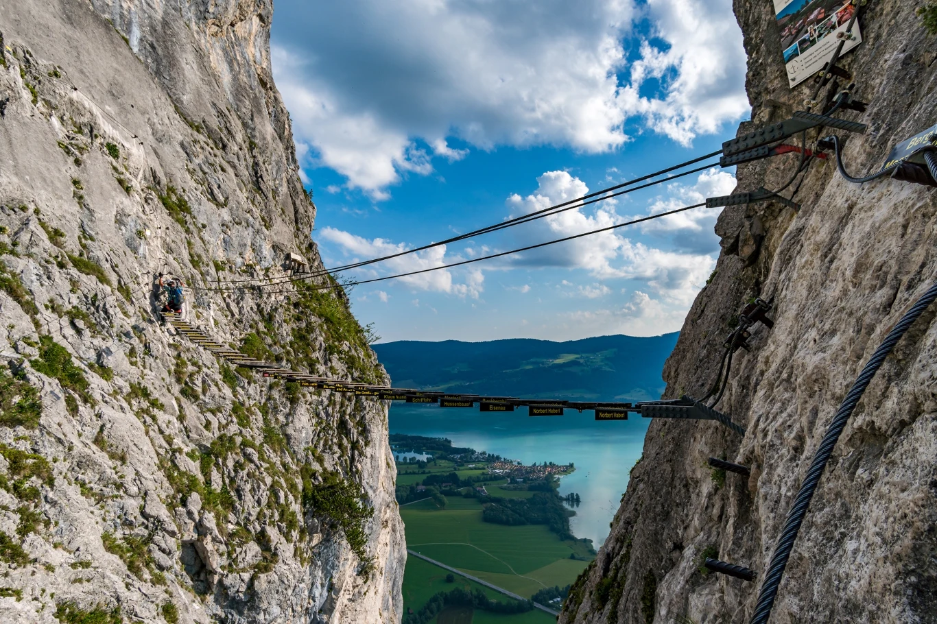 Via Ferrata - Drachenwand w Austrii Wąska metalowa kładka zawieszona wysoko pomiędzy stromymi skałami z widokiem na jezioro i zieloną dolinę w tle oraz fragmentami nieba z chmurami.